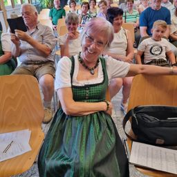 Eine Frau in einem grünen Dirndl lächelt, während sie in einem Stuhl in einem Raum mit anderen Menschen sitzt. Ein Junge sitzt neben ihr.