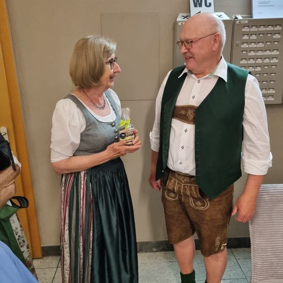 Ein älteres Paar, gekleidet in traditioneller bayerischer Kleidung, steht lächelnd zusammen. Die Frau hält ein verpacktes Geschenk. Hinter ihnen befindet sich eine Wand mit einem WC-Schild.