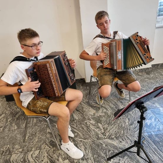Zwei Jungen sitzen und spielen jeweils ein Akkordeon. Der Junge auf der linken Seite trägt ein weißes T-Shirt, eine Brille und Shorts. Der Junge auf der rechten Seite trägt ein weißes T-Shirt und Shorts mit einem Logo. Sie befinden sich in einem gefliesten Raum.