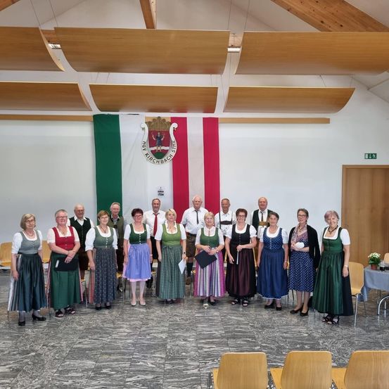 Eine Gruppe von Erwachsenen in traditioneller bayerischer Kleidung steht in einem Raum mit weißer Wand, einer Flagge und einem Logo. Einige halten Dokumente. Der Raum hat Holzstühle und eine Holztür.
