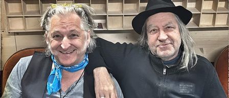 Zwei ältere Männer sitzen lächelnd zusammen, einer trägt eine blaue Schürze und eine schwarze Halskette, der andere einen schwarzen Hut und einen Ring. Sie stehen vor einem Holzregal.