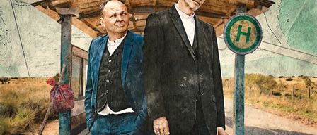 Zwei Männer in formeller Kleidung stehen an einer Bushaltestelle. Einer trägt einen blauen Anzug, der andere einen schwarzen. Sie lächeln und schauen in die Kamera. Hinter ihnen ist eine Karte und ein Schild mit dem Buchstaben 'H'.