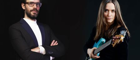 Zwei Personen, ein Mann und eine Frau, stehen vor einem dunklen Hintergrund. Der Mann trägt eine Brille und einen schwarzen Anzug und hat die Arme verschränkt. Die Frau hält eine blaue E-Gitarre.