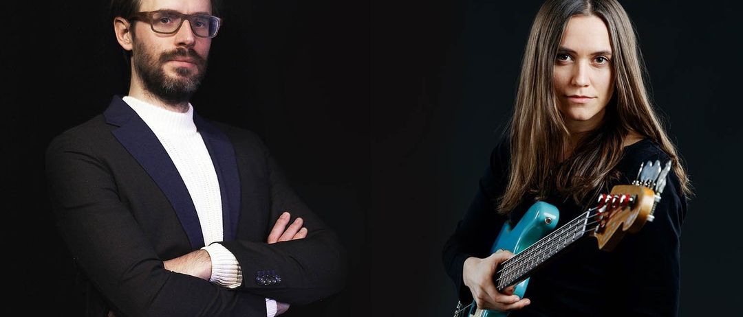 Zwei Personen, ein Mann und eine Frau, stehen vor einem dunklen Hintergrund. Der Mann trägt eine Brille und einen schwarzen Anzug und hat die Arme verschränkt. Die Frau hält eine blaue E-Gitarre.