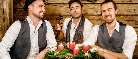 Drei Männer lächeln und posieren für ein Foto vor einer Holzwand mit einer Weihnachtsgirlande und Kerzen. Ein Mann hält eine Geige.