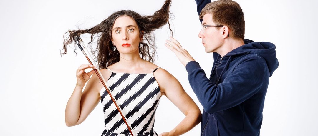 Eine Frau mit lockigem Haar hält einen Bogen und einen Mann in einer blauen Kapuzenjacke, der ihr Haar mit einer Klammer hält.