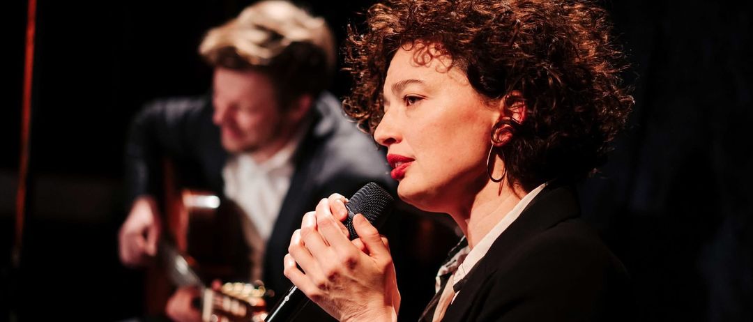 Eine Frau mit lockigen Haaren singt in ein Mikrofon, während ein Mann im Hintergrund Gitarre spielt.