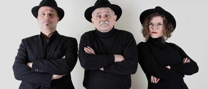 Drei Menschen in schwarzen Kleidungsstücken und Hüten stehen in einer Reihe vor einem weißen Hintergrund. Der Mann auf der linken Seite hat die Arme verschränkt. Der Mann in der Mitte hat einen Schnurrbart und faltet die Arme. Die Frau auf der rechten Seite trägt eine Brille und hat die Arme verschränkt.