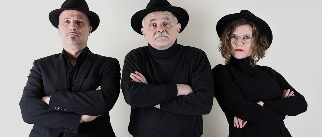 Drei Menschen in schwarzen Kleidungsstücken und Hüten stehen in einer Reihe vor einem weißen Hintergrund. Der Mann auf der linken Seite hat die Arme verschränkt. Der Mann in der Mitte hat einen Schnurrbart und faltet die Arme. Die Frau auf der rechten Seite trägt eine Brille und hat die Arme verschränkt.