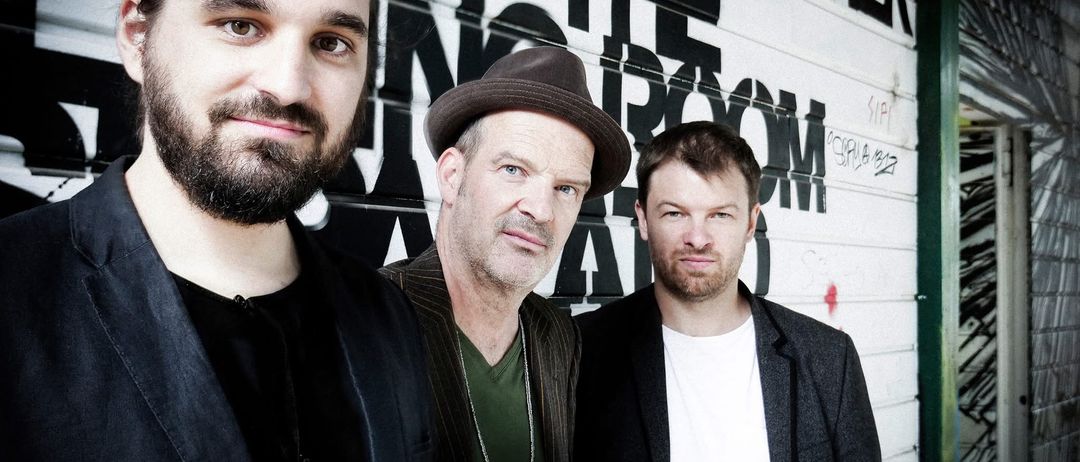 Drei Männer posieren für ein Foto vor einer Graffitiwand mit 'AT THE ROOM' und 'SIP'. Der Mann links hat einen Bart und eine schwarze Jacke.