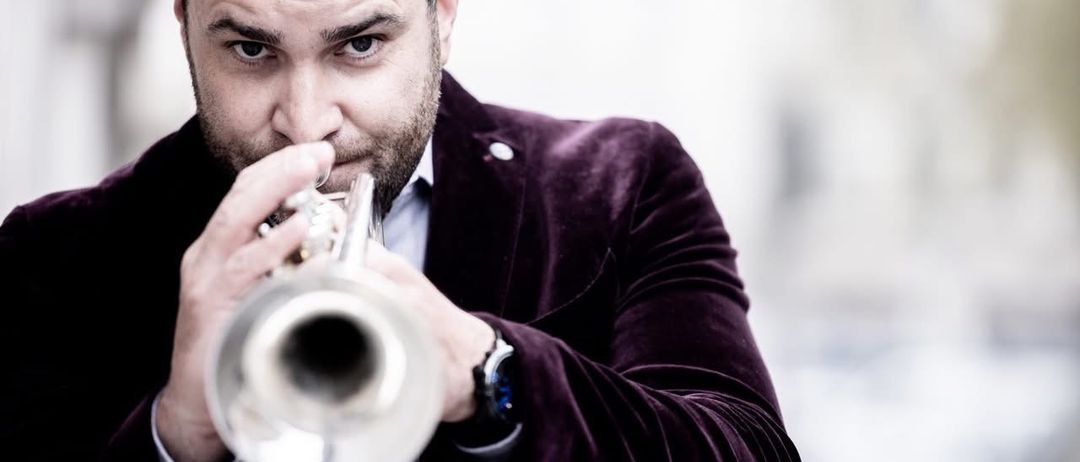 Ein Mann spielt mit ernster Miene eine Trompete und trägt einen lila Anzug und eine Uhr.