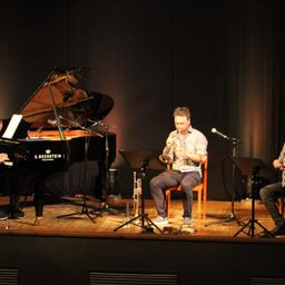 Ein Pianist spielt auf einem schwarzen Flügel, begleitet von einem Trompeter und einem Klarinettisten auf der Bühne.
