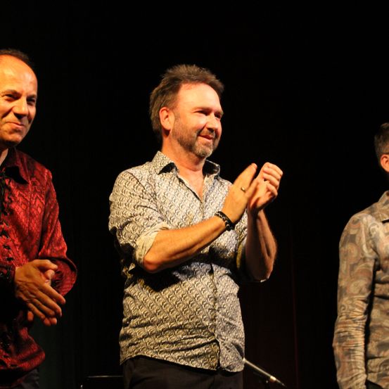 Drei Männer stehen lächelnd auf der Bühne. Der mittlere klatscht. Der linke trägt ein rotes Shirt, der rechte ein Camouflage-Shirt.