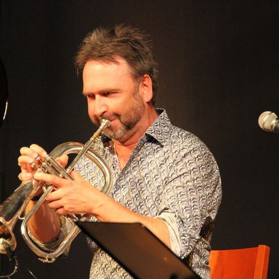 Ein Mann spielt Trompete auf der Bühne. Er trägt ein gemustertes Hemd und hat ein Mikrofon vor sich.