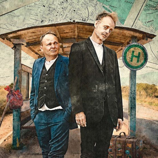 Zwei Männer stehen an einem Bahnhof, einer trägt einen blauen Anzug, der andere einen schwarzen. Sie stehen neben einem Gepäckwagen. Dahinter befindet sich eine Karte auf einem hölzernen Unterstand mit einem H-Schild.