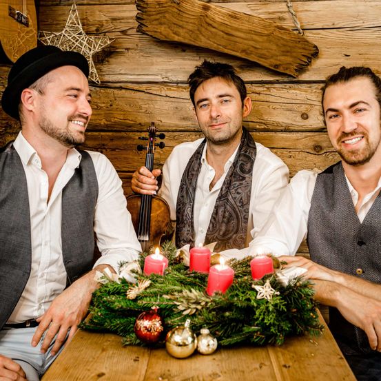 Drei Männer sitzen um einen Tisch mit einem Weihnachtskranz und Kerzen. Einer hält eine Geige. Sie tragen Westen und weiße Hemden.