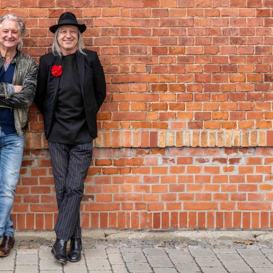 Zwei ältere Männer stehen neben einer Ziegelmauer und lächeln. Einer trägt einen schwarzen Hut und eine schwarze Jacke, der andere eine Lederjacke.