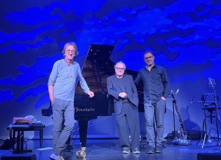 Drei Männer stehen auf einer Bühne mit Musikinstrumenten vor einem blauen Hintergrund. Ein Mann hält einen Flügel.