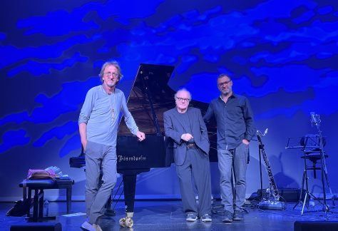 Drei Männer stehen auf einer Bühne mit Musikinstrumenten vor einem blauen Hintergrund. Ein Mann hält einen Flügel.