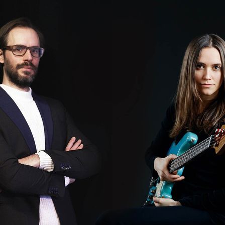 Zwei Personen stehen vor einem schwarzen Hintergrund. Der Mann, der eine Brille und einen Anzug trägt, hat die Arme verschränkt. Die Frau, die ein schwarzes Oberteil trägt, spielt eine türkisfarbene Bassgitarre.