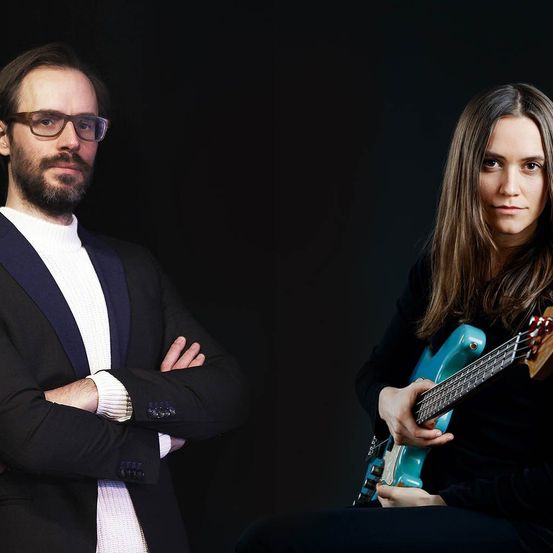 Zwei Personen stehen vor einem schwarzen Hintergrund. Der Mann, der eine Brille und einen Anzug trägt, hat die Arme verschränkt. Die Frau, die ein schwarzes Oberteil trägt, spielt eine türkisfarbene Bassgitarre.