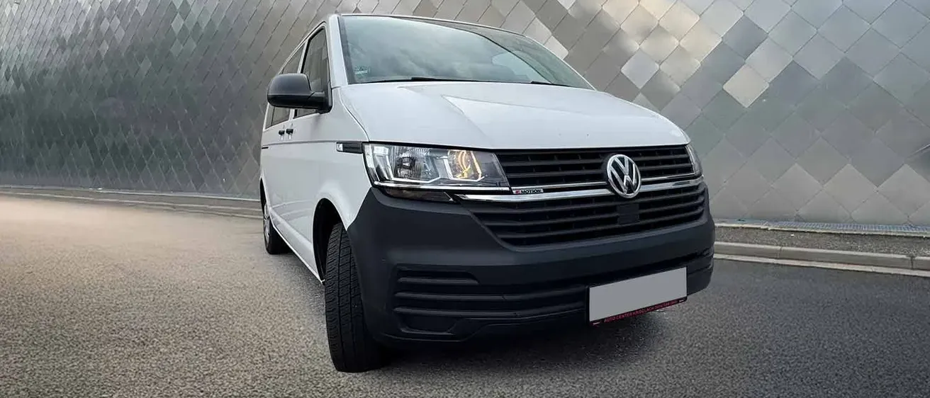 Ein weißer Volkswagen-Transporter mit schwarzem Stoßfänger steht vor einem Gebäude mit einer rautenförmigen Wand.