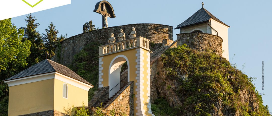 Eine Steinburg auf einem Hügel mit Statuen, einem Kreuz und einer steilen Treppe. Das Gebäude hat gelbe Wände und grüne Pflanzen an der Seite.