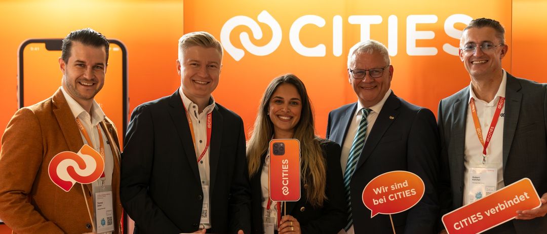 Fünf Personen posieren für ein Foto an einem Stand mit orangefarbenem Hintergrund. Sie halten Schilder mit der Aufschrift 'CITIES'. Eine Frau hält ein Handy. Dahinter steht ein größeres Schild mit der Aufschrift 'CITIES'.
