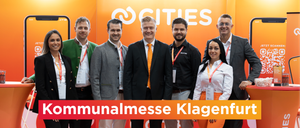 Five people stand smiling in front of an orange banner with the word Cities written in white. The people are wearing ID badges.