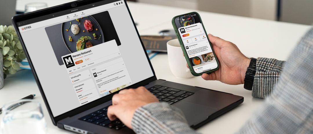 Eine Person benutzt einen Laptop und hält ein Smartphone, beide zeigen eine Restaurant-Website an. Der Laptop-Bildschirm zeigt eine detaillierte Restaurant-Seite mit Fotos und Text. Der Smartphone-Bildschirm zeigt eine Restaurant-App mit einem Menü.