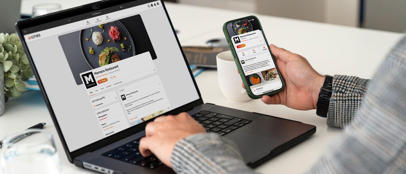 Eine Person benutzt einen Laptop und hält ein Smartphone, beide zeigen eine Restaurant-Website an. Der Laptop-Bildschirm zeigt eine detaillierte Restaurant-Seite mit Fotos und Text. Der Smartphone-Bildschirm zeigt eine Restaurant-App mit einem Menü.
