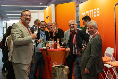 Eine Gruppe von Männern steht bei einem Event um einen Tisch herum, lächelt und hält Weingläser. Sie tragen formelle Kleidung, und der Tisch hat Flaschen und einen Regenschirm. Orange Wände mit dem Wort 'cities' im Hintergrund.