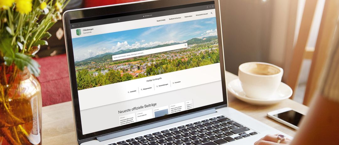 Ein Laptop auf einem Schreibtisch zeigt eine Website mit 'Neueste offizielle Beiträge'. In der Nähe sind eine Kaffeetasse, ein Telefon und eine Vase mit Blumen.