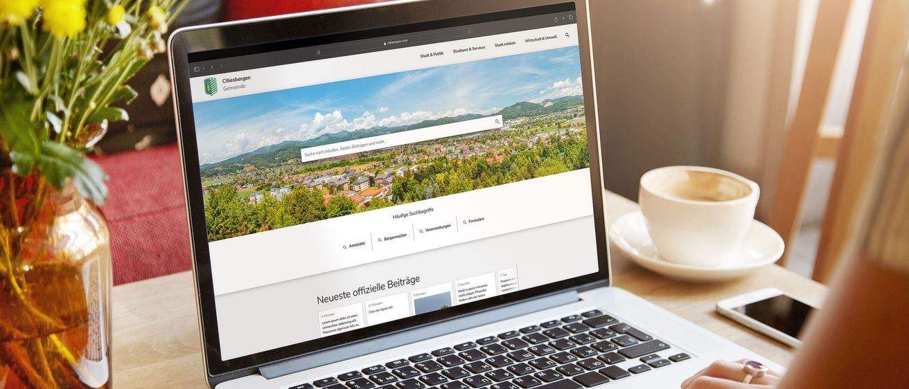 Ein Laptop auf einem Schreibtisch zeigt eine Website mit 'Neueste offizielle Beiträge'. In der Nähe sind eine Kaffeetasse, ein Telefon und eine Vase mit Blumen.