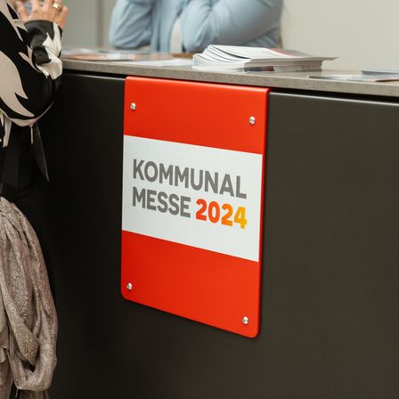 Ein orangefarbenes Schild mit weißem Text, das 'KOMMUNAL MESSE 2024' steht, ist auf einer grauen Oberfläche montiert. Eine Person steht in der Nähe.