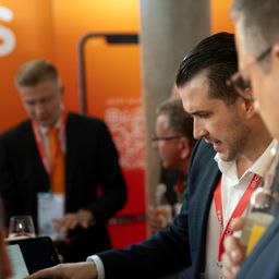 Drei Männer in Geschäftskleidung bei einer Veranstaltung, einer zeigt auf ein Tablet, einer trinkt aus einem Glas, ein anderer mit einem roten Ausweisband.