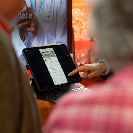 Eine Person interagiert mit einem Tablet und zeigt auf eine Karte auf dem Bildschirm. Dahinter hält eine Person ein Smartphone mit 'zielle News' sichtbar.