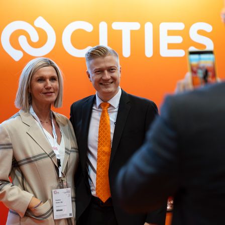 Zwei Personen, eine Frau und ein Mann, stehen nebeneinander und lächeln für ein Foto vor einem Hintergrund mit dem Wort 'CITIES'. Die Frau trägt einen beigen Blazer, und der Mann hat eine orange Krawatte. Hinter ihnen hält eine Person ein Handy.