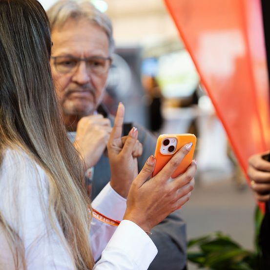 Eine Frau mit langen Haaren und einer orangefarbenen iPhone-Hülle spricht mit einem Mann im Anzug, der mit den Fingern gestikuliert.