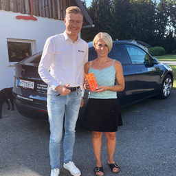 Ein Mann und eine Frau stehen vor einem schwarzen Auto und halten eine Geschenkbox. Der Mann trägt ein weißes Hemd und Jeans, die Frau ein ärmelloses Top und einen Rock. Das Kennzeichen des Autos lautet 'W 12695 N'.