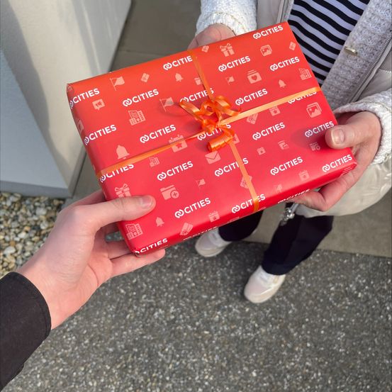 Zwei Personen halten eine rote Geschenkbox, die mit rotem Papier und weißen Symbolen und Text verpackt ist. Eine Person trägt eine weiße Jacke, die andere ein schwarzes Hemd.