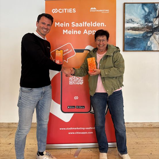 Zwei Personen stehen vor einem orangefarbenen Banner mit der Aufschrift Mein Saalfelden. Meine App. Sie halten orangefarbene Schachteln mit einem QR-Code.