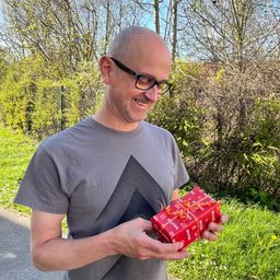 Ein Mann mit Brille lächelt, während er ein rotes Geschenk mit einem Band im Freien hält.