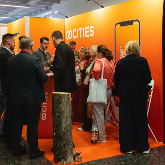 Menschen sind an einem Stand versammelt, möglicherweise bei einer Veranstaltung. Einige halten Gläser, und eine Frau hält eine Handtasche. Eine orangefarbene Wand mit dem Text '@CITIES' steht hinter ihnen.