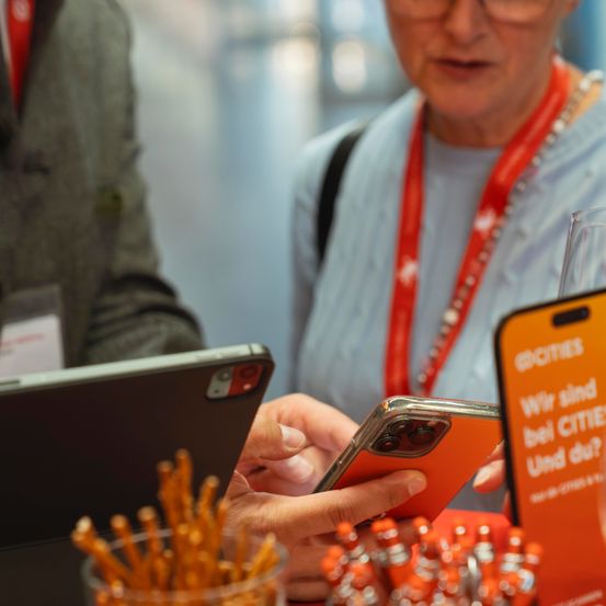 Eine ältere Frau hält ein oranges Smartphone, möglicherweise in einem Konferenzsaal. Eine andere Person hält den Bildschirm des Telefons mit den Händen. Ein Glas steht neben einem Telefon.