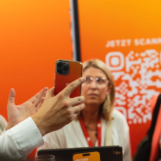 Eine Person hält ein Handy vor einer Frau mit Brille. Das Handy hat einen roten Rücken und eine schwarze Kamera. Im Hintergrund befindet sich eine orangefarbene Wand mit einem QR-Code und den Worten 'JETZT SCAN'.