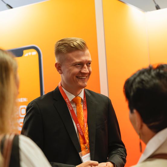 Ein Mann mit Namensschild lächelt und unterhält sich mit zwei Personen vor einer orangefarbenen Wand.