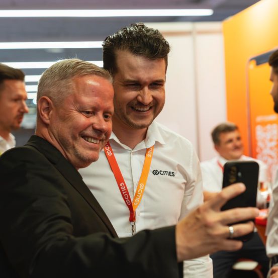 Zwei Männer, beide in weißen Hemden, machen ein Selfie mit einem Telefon. Der Mann vorn trägt einen schwarzen Anzug. Im Hintergrund sind andere Männer, möglicherweise Kollegen.