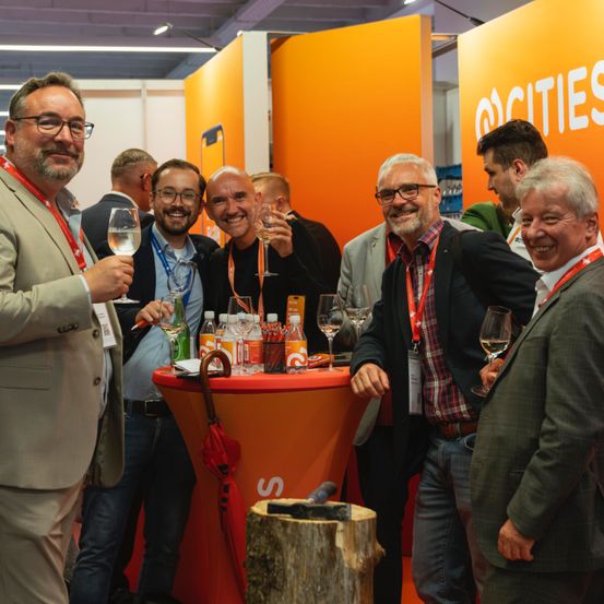 Eine Gruppe von Männern bei einer Messe, die mit Weinläsern vor einem orangefarbenen Stand mit dem Text 'cities' anstoßen.
