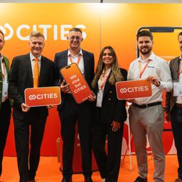 Fünf Personen stehen in einer Reihe und halten orange Schilder mit der Aufschrift 'Cities'. Dahinter ist ein Banner mit demselben Wort und einem Smartphone-Bild.